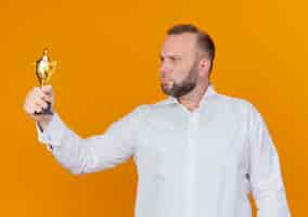 Foto gratuita hombre barbudo con camisa blanca sosteniendo el trofeo mirándolo con cara seria de pie sobre la pared naranja