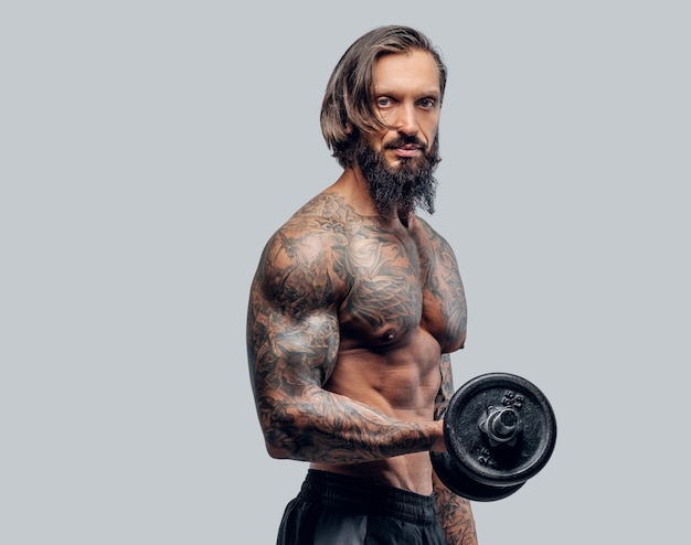 Hombre barbudo sin camisa con cuerpo tatuado sostiene pesa sobre fondo gris.