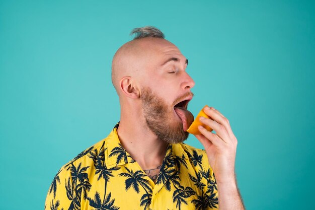 Un hombre barbudo con una camisa con un estampado de palmeras en una pared turquesa lame una naranja