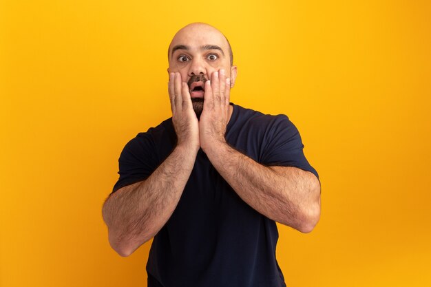 Hombre barbudo en camiseta azul marino asombrado y preocupado con las manos en la cara de pie sobre la pared naranja