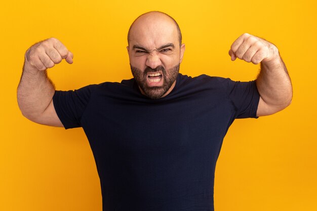 Hombre barbudo en camiseta azul marino con cara enojada levantando puños loco loco parado sobre pared naranja