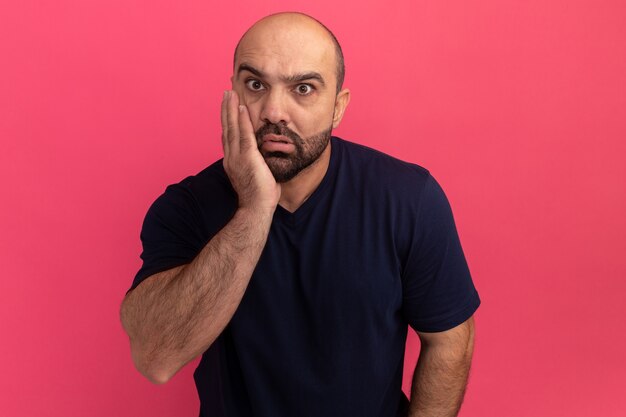 Hombre barbudo en camiseta azul marino confundido y preocupado con el brazo en la mejilla de pie sobre la pared rosa