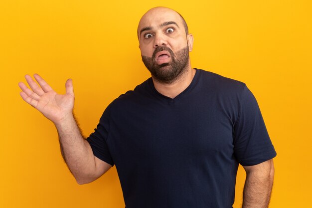 Hombre barbudo en camiseta azul marino confundido presentando algo con el brazo de él tenía parado sobre la pared naranja