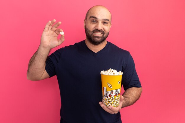 Hombre barbudo en camiseta azul marino cubo con palomitas de maíz con sonrisa en la cara sosteniendo de pie sobre la pared rosa