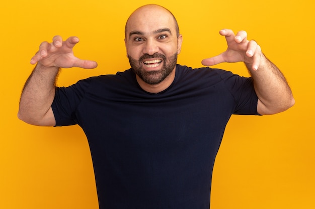 Hombre barbudo en camiseta azul marino feliz y alegre haciendo gestos de garras como un oso parado sobre una pared naranja