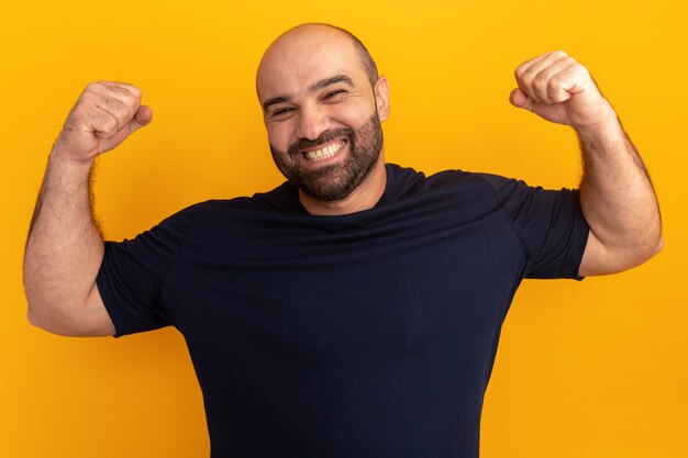 Hombre barbudo en camiseta azul marino feliz y emocionado levantando los puños como un ganador de pie sobre una pared naranja