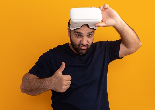 Hombre barbudo en camiseta azul marino con gafas de realidad virtual sonriendo mostrando los pulgares para arriba de pie sobre la pared naranja