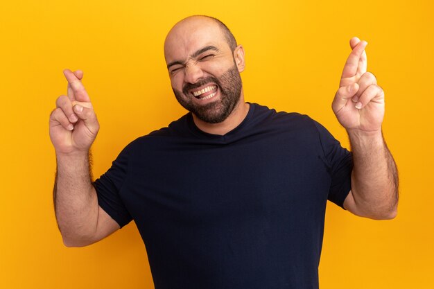 Hombre barbudo en camiseta azul marino haciendo deseo deseable con expresión de esperanza cruzando los dedos con los ojos cerrados de pie sobre la pared naranja