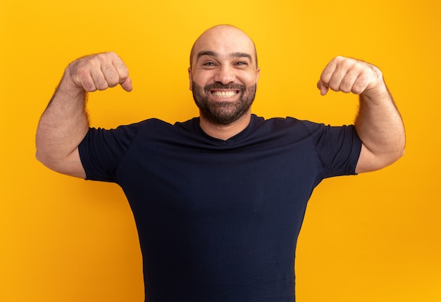 Hombre barbudo en camiseta azul marino levantando los puños como un ganador feliz y emocionado sonriendo ampliamente de pie sobre la pared naranja