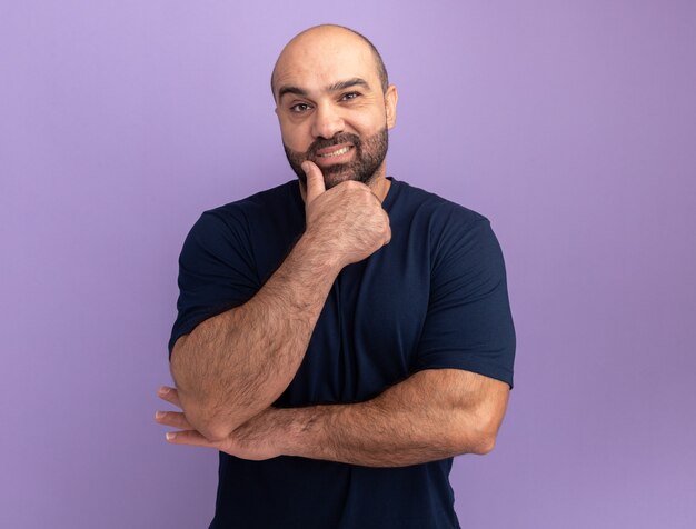 Hombre barbudo en camiseta azul marino con la mano en el ching sonriendo confiado de pie sobre la pared púrpura