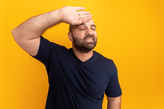 Hombre barbudo en camiseta azul marino con la mano en la frente con expresión molesta de pie sobre la pared naranja