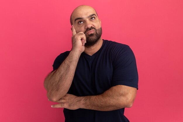 Hombre barbudo en camiseta azul marino mirando hacia arriba desconcertado parado sobre pared rosa