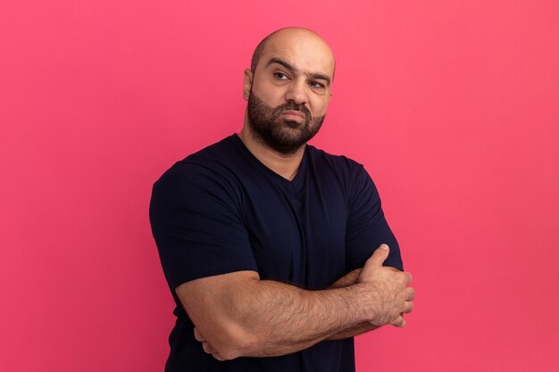 Hombre barbudo en camiseta azul marino mirando asdie con los brazos cruzados disgustado de pie sobre la pared rosa