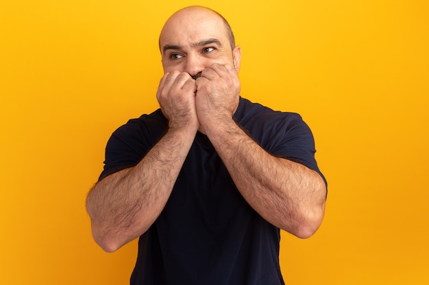 Hombre barbudo en camiseta azul marino mirando a un lado estresado y nervioso morderse las uñas de pie sobre la pared naranja