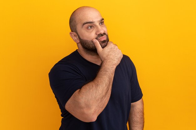 Hombre barbudo en camiseta azul marino mirando a un lado con expresión pensativa con la mano en la barbilla de pie sobre la pared naranja