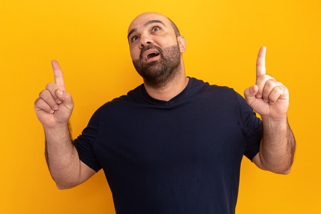 Hombre barbudo en camiseta azul marino mirando sorprendido apuntando con los dedos índices de pie sobre la pared naranja