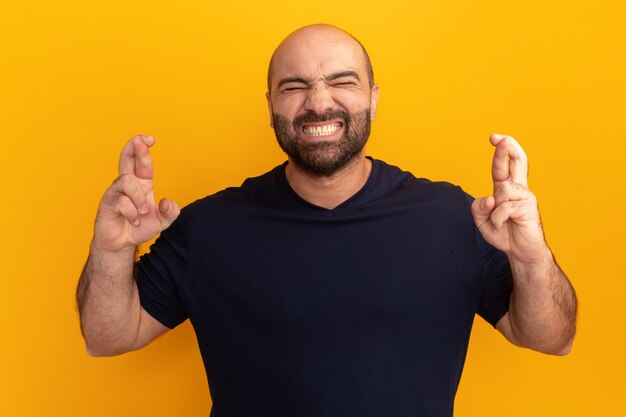 Hombre barbudo en camiseta azul marino con los ojos cerrados haciendo deseo cruzar los dedos de pie sobre la pared naranja