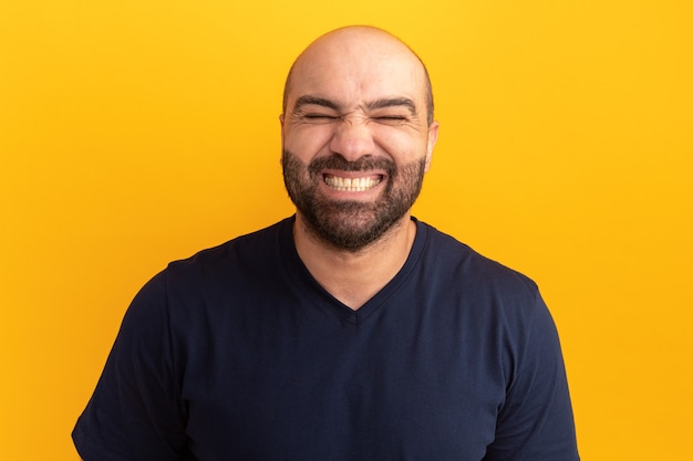 Hombre barbudo en camiseta azul marino con los ojos cerrados sonriendo alegremente mostrando los dientes de pie sobre la pared naranja