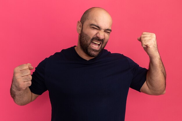 Hombre barbudo en camiseta azul marino de pie con los ojos cerrados apretando los puños pared rosa feliz y emocionada