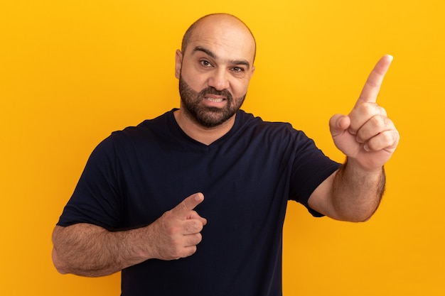 Foto gratuita hombre barbudo en camiseta azul marino que parece confundido y disgustado apuntando con el dedo índice a algo parado sobre la pared naranja