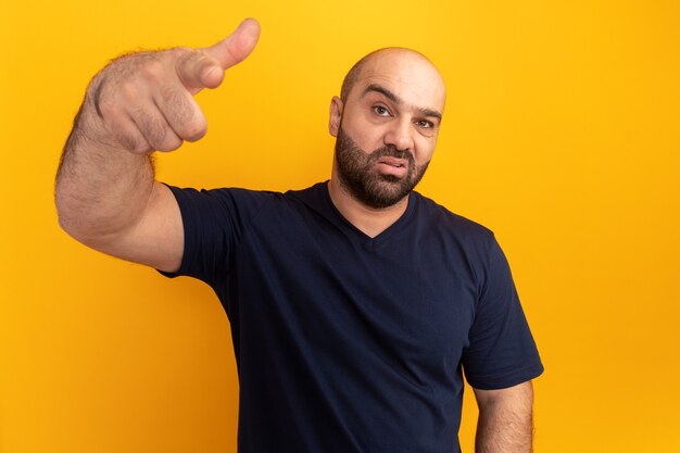 Hombre barbudo en camiseta azul marino que parece confundido y muy decepcionado apuntando con el dedo índice sobre la pared naranja