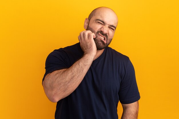 Hombre barbudo en camiseta azul marino que parece confundido rascándose la cara de pie sobre la pared naranja