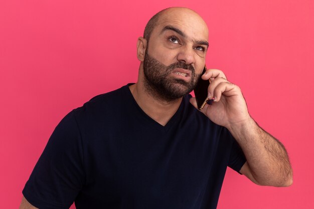 Hombre barbudo en camiseta azul marino que parece molesto e irritado mientras habla por teléfono móvil de pie sobre una pared rosa