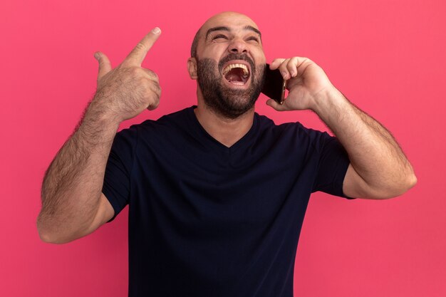 Hombre barbudo en camiseta azul marino que parece molesto gritando volviéndose loco mientras habla por teléfono móvil de pie sobre la pared rosa