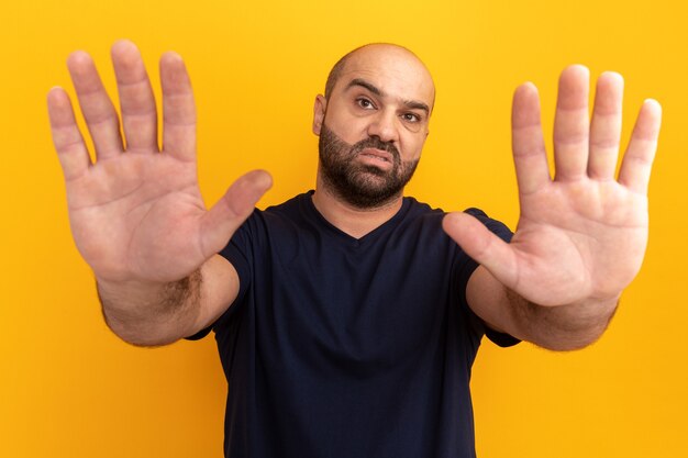 Hombre barbudo en camiseta azul marino con rostro serio haciendo gesto de parada con las manos de pie sobre la pared naranja