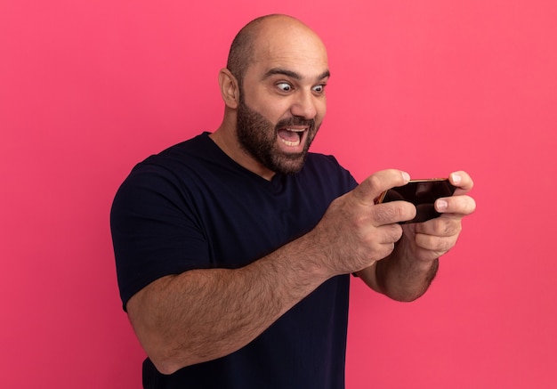 Hombre barbudo en camiseta azul marino con smartphone jugando juegos mirando sorprendido y emocionado de pie sobre la pared rosa