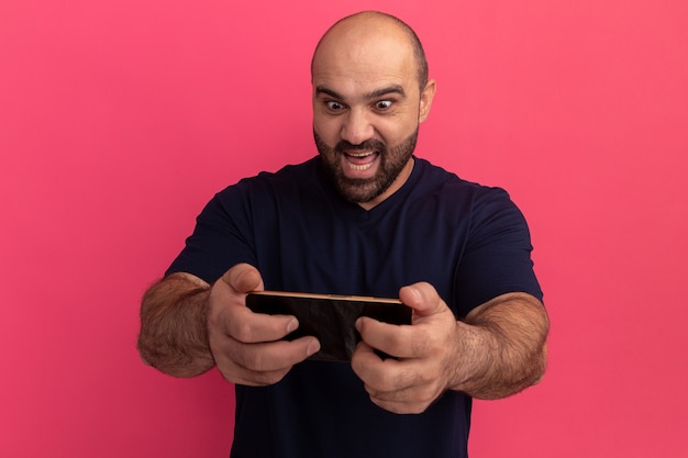 Hombre barbudo en camiseta azul marino con smartphone jugando juegos de pie feliz y emocional sobre pared rosa