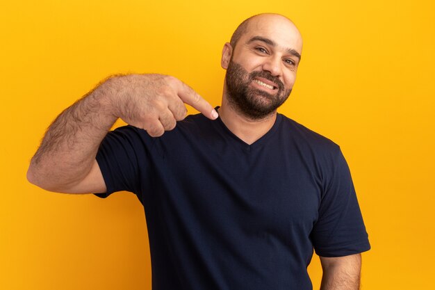 Hombre barbudo en camiseta azul marino sonriendo confiado apuntando con el dedo índice a sí mismo de pie sobre la pared naranja