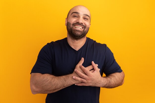 Hombre barbudo en camiseta azul marino sonriendo con las manos en el pecho sintiéndose agradecido de pie sobre la pared naranja