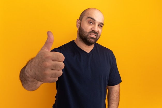 Hombre barbudo en camiseta azul marino sonriendo mostrando los pulgares para arriba de pie sobre la pared naranja