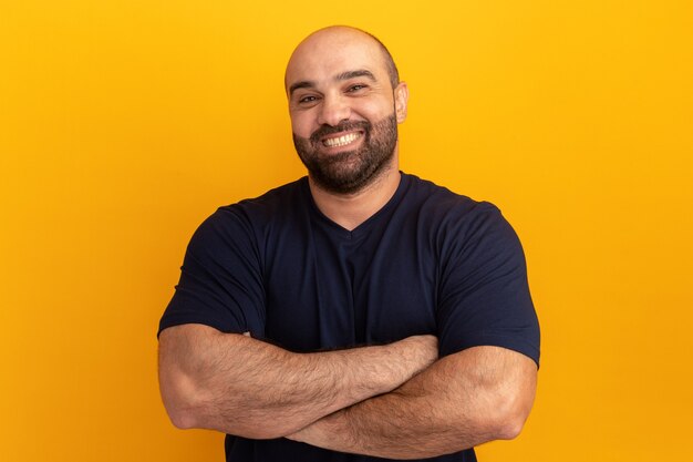 Hombre barbudo en camiseta azul marino con sonrisa en la cara con los brazos cruzados de pie sobre la pared naranja