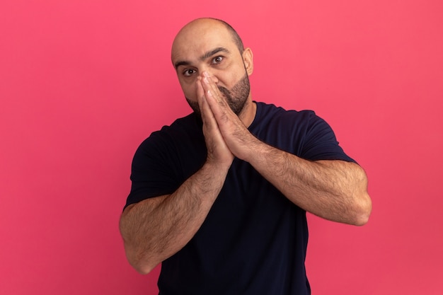 Hombre barbudo en camiseta azul marino sosteniendo las palmas juntas frente a su rostro estresado y preocupado de pie sobre la pared rosa