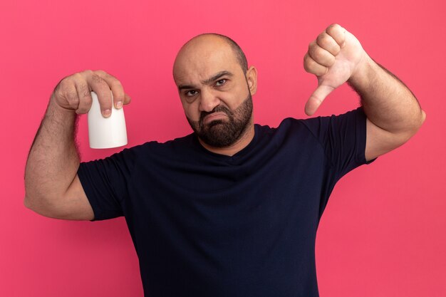 Hombre barbudo en camiseta azul marino sosteniendo una taza disgustado mostrando los pulgares hacia abajo de pie sobre la pared rosa