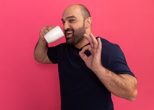 Hombre barbudo en camiseta azul marino sosteniendo una taza de té va a beber feliz y positivo mostrando el signo de ok de pie sobre la pared rosa