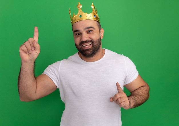 Hombre barbudo en camiseta blanca con corona de oro feliz y positivo apuntando con los dedos índices de pie sobre la pared verde