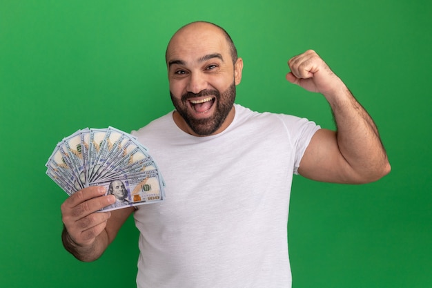 Hombre barbudo en camiseta blanca con dinero en efectivo feliz y emocionado levantando el puño de pie sobre la pared verde