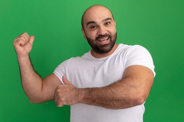 Hombre barbudo en camiseta blanca feliz y alegre apuntando con los dedos hacia atrás de pie sobre la pared verde