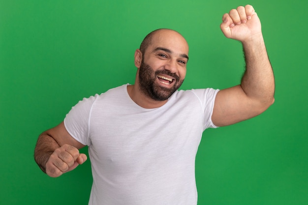 Hombre barbudo en camiseta blanca feliz y emocionado levantando los puños de pie sobre la pared verde