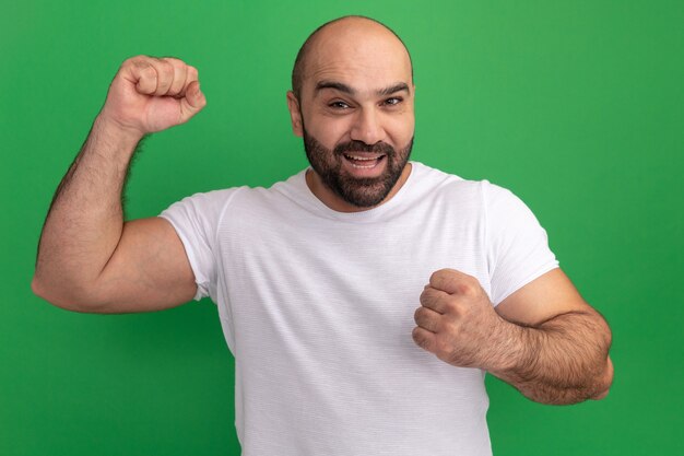 Hombre barbudo en camiseta blanca feliz y emocionado levantando los puños de pie sobre la pared verde