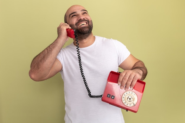 Foto gratuita hombre barbudo en camiseta blanca hablando por un teléfono antiguo sonriendo con cara feliz de pie sobre la pared verde