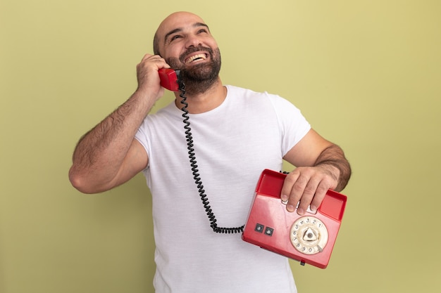 Hombre barbudo en camiseta blanca hablando por un teléfono antiguo sonriendo con cara feliz de pie sobre la pared verde