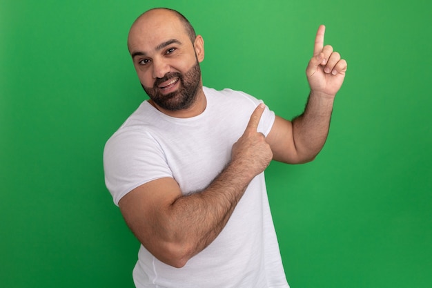 Hombre barbudo en camiseta blanca con smie en la cara apuntando con el dedo índice hacia el lado sonriendo alegremente de pie sobre la pared verde