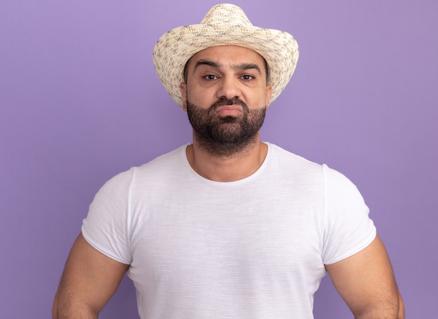 Hombre barbudo en camiseta blanca y sombrero de verano con expresión seria y segura de pie sobre la pared púrpura