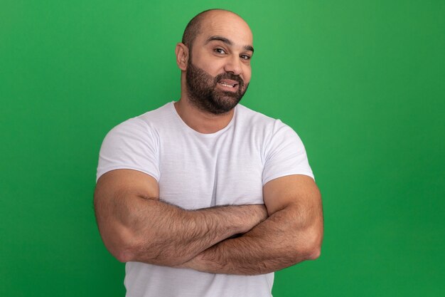 Hombre barbudo en camiseta blanca con sonrisa en la cara con los brazos cruzados de pie sobre la pared verde