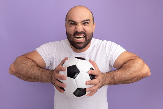 Hombre barbudo en camiseta blanca sosteniendo un balón de fútbol con cara enojada gritando volviéndose loco de pie sobre la pared púrpura