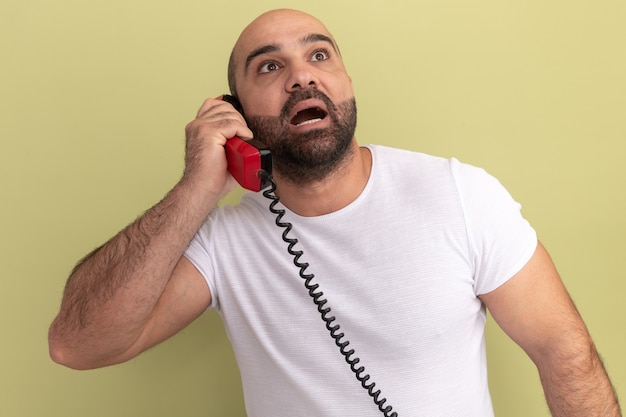 Hombre barbudo en camiseta blanca sosteniendo teléfono antiguo mirando asombrado y sorprendido de pie sobre la pared verde